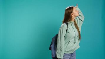 Curious smiling girl holding rucksack looking around for something, doing hand gesturing. Upbeat person doing surveillance gesture, exploring, seeking things, isolated over studio backdrop, camera B photo