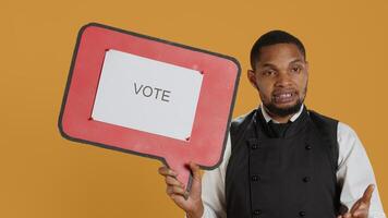 Professional waiter showing a message to vote on speech bubble, creating an advertisement for politics on cardboard icon. Restaurant employee working in customer oriented industry. Camera A. photo