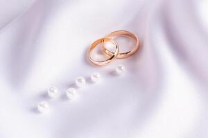 A pair of gold wedding rings on a white satin background with a scattering of several pearls. postcard. invitation. A copy space. minimalism. photo