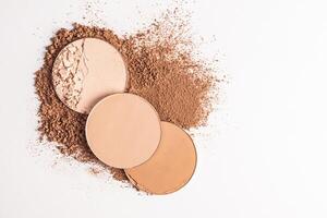 Three round blocks of compact powder in various tones lie on a sample of loose powder in a bronze tone. top view. white background. A copy space. photo