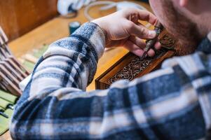 man artisan carpenter carves pattern on wooden casket with a chisel in workshop photo