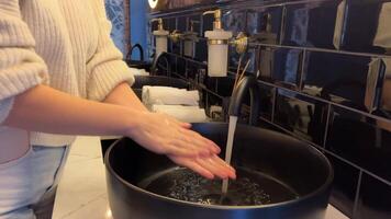 hand touches the water and washes hands with soap. A black sink with a silver faucet next to an oval mirror and a shelf with hand paper towels. Close-up video