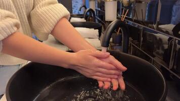 Washing hands in a black sink. black toilet is in a bathroom with a black tile wall. hand touches the water and washes sink next to an oval mirror and a shelf with hand paper towels. Close-up video