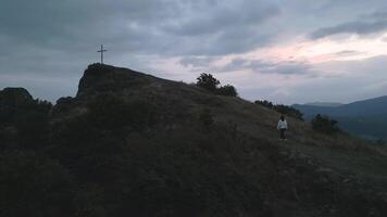 Aerial panning view caucasian woman in white walk uphill to mountain hilltop with giant standing cross on dramatic sunset and cloudy evening in nature. Believer search for purpose hope conceptual video