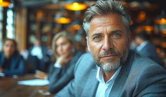 A man in a gray suit is sitting at a table with other people. He is looking at the camera and he is focused photo