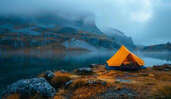 A yellow tent is set up on a rocky shore near a lake. The sky is cloudy and the water is calm photo