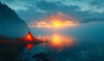 A small teepee is on the shore of a lake. The sun is setting, casting a warm glow over the scene. photo