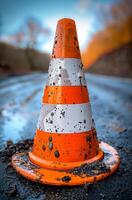 A dirty orange and white cone is on the road. The cone is in the middle of the road and is surrounded by mud photo
