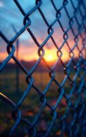 A fence with a chain link pattern and a sun shining through it. The sun is setting and the sky is a beautiful orange color photo