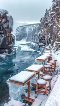 A long row of tables and stools blanketed in snow beside a river photo