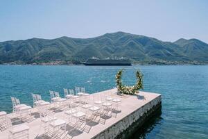 White chairs stand in front of a round wedding arch by the sea, with a cruise ship in the background photo