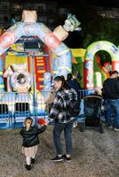 Mother and a little girl stand holding hands in front of a colorful attraction. Back view photo