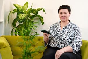 Successful and goal-oriented middle-aged woman demonstrating business proficiency while talking on the phone while sitting comfortably on a sofa in a modern office photo