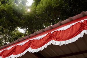 Red and white flag decoration hanging on building roof isolated on horizontal outdoor green tree leaves and bright sky background. Bendera merah putih fabric cloth texture. photo