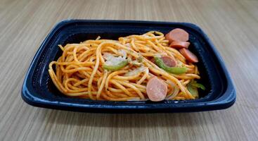 Cheap convinience store spaghetti with tomato sauce and sausage meal in black plastic plate container isolated on horizontal wooden table surface background. photo