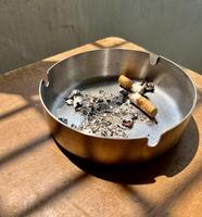 Round metal aluminium ash tray with cigarette waste and ashes isolated on vertical wooden surface background with window shadow from sun lights. photo