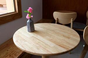 Bright pink artificial sakura flower in dark gray vase on light brown round wood table beside window. photo