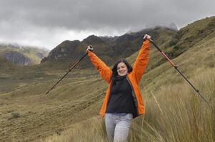 woman in red jacket raising her arms with trekking poles in the middle of the moor with mountains in the background on a cloudy day photo