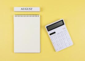 flat layout of opened blank page notebook with wooden calendar August and white calculator on yellow background. monthly financial plan concept. photo