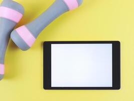 flat lay of digital tablet with blank white screen and two pink grey dumbbells isolated on yellow background. photo
