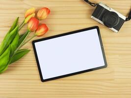 flat lay of digital tablet with blank white screen, tulip flowers and digital camera isolated on wooden table background. photo