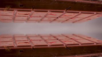 Close-up a milk ruby chocolate bar with chunks of nuts almonds and strawberry, displayed against a gray background is placed on a reflective surface, creating a mirrored image of itself video
