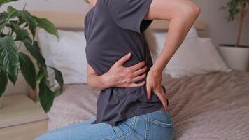 Close-up of a woman grabbing with her hands the place of pain in the lower back and cross while sitting on the bed. Joint pain. Menstrual pain, cramping. video