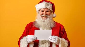 Santa Claus holding blank white card in front of Christmas tree and yellow background. Concept of festive holiday season, Christmas greetings. Greeting card, postcard, mockup, invitation. Copy space photo