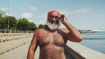 Muscular older man with a gray beard and red swim cap standing on a sunny waterfront promenade, adjusting his sunglasses, smiling confidently, green trees and a clear blue sky in the background video