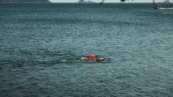 Open water swimmer in a large lake with orange safety buoy and swim cap, performing a freestyle stroke, calm and expansive water surface under a partly cloudy sky, distant motorboat visible, outdoor video