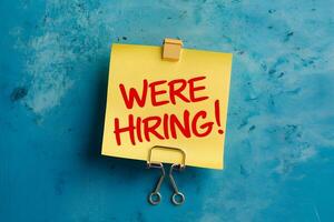 Blue background, yellow post it note with Were Hiring in red, metallic clip photo