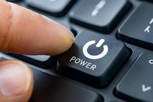Finger presses black power button on keyboard, white power icon, blurred keys photo