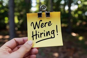 Hand holding yellow sticky note with Were Hiring against blurred trees photo