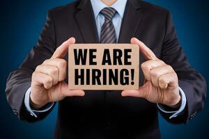 Man in suit holding WE ARE HIRING card, dark blue background photo