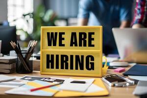 Office desk with We Are Hiring sign, office supplies, laptop, blurred figure photo
