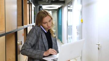 A woman in business attire manages multiple tasks in an office hallway. Professional balancing a laptop and phone call simultaneously. video