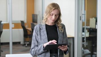 A businesswoman in smart casual attire demonstrates comfort with digital tools in the workplace. Woman standing in front of her desk using a digital device, looking at camera with a smile. video