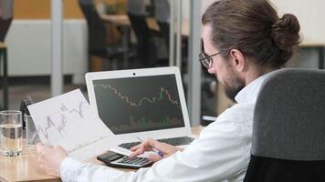 Man with glasses is seated at his desk, analyzing financial charts on his laptop. Professional office worker closely examines market data while holding a printout of graphs and calculating video