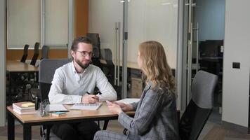 Two professionals engaged in a one-on-one meeting in an office setting. A man with glasses and beard taking notes while a woman gestures as she speaks. video