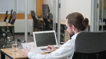 A man with glasses is seated at his desk, analyzing financial charts on his laptop. Businessman an office setting reviews statistical data on a laptop and paper charts. video