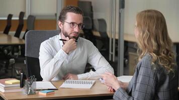 Two professionals sitting on a shared table inside a modern office. Businessman take notes and touch his chin while listening to his workmate explaining about work while holding a mobile phone. video