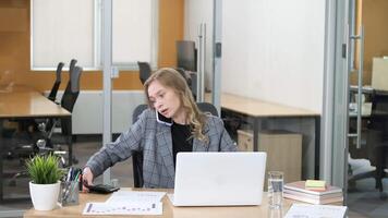 professionell kvinna talande på henne smartphone medan arbetssätt på henne bärbar dator, beräknande och granskning data rapporterar. kontor arbetstagare håller på med göra flera saker samtidigt i en modern kontor. video