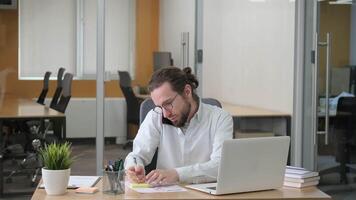 A businessman doing a multi-tasking job on his desk at office. He is busy discussing a marketing reports on his hands through cellphone while talking notes at the same time working on his laptop. video