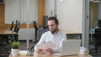 Concentrated businessman on eyeglasses checking and comparing each marketing report, analyzing statistics data in charts. Man working on computer alone in modern office. video