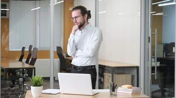 An office worker standing and walking against at his desk while hold a pen and touching his chin. A man with glasses serious working on his laptop. Paperwork and books on tidy desk. video