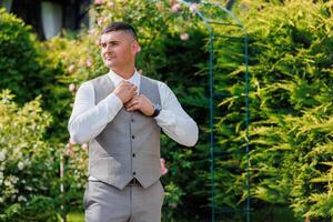 A man in a suit and vest stands in front of a green archway. He is adjusting his tie and he is in a professional setting photo