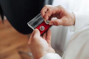 A man is holding a pair of earrings in a red box. The earrings are silver and have a design on them. The man is wearing a white shirt and he is getting ready for a special occasion photo