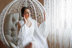 A woman in a white gown sits in a white chair. She is looking out the window photo