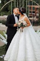 A bride and groom are kissing in front of a wedding arch. The bride is holding a bouquet of flowers photo