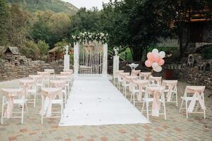 A wedding ceremony is taking place in a garden with a white archway and pink balloons. The chairs are arranged in rows, and the guests are seated on them. The atmosphere is elegant and romantic photo